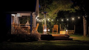 Patio esterno con arredi in legno, un tavolo e sedie, illuminato da una ghirlanda di luci e un lampione.