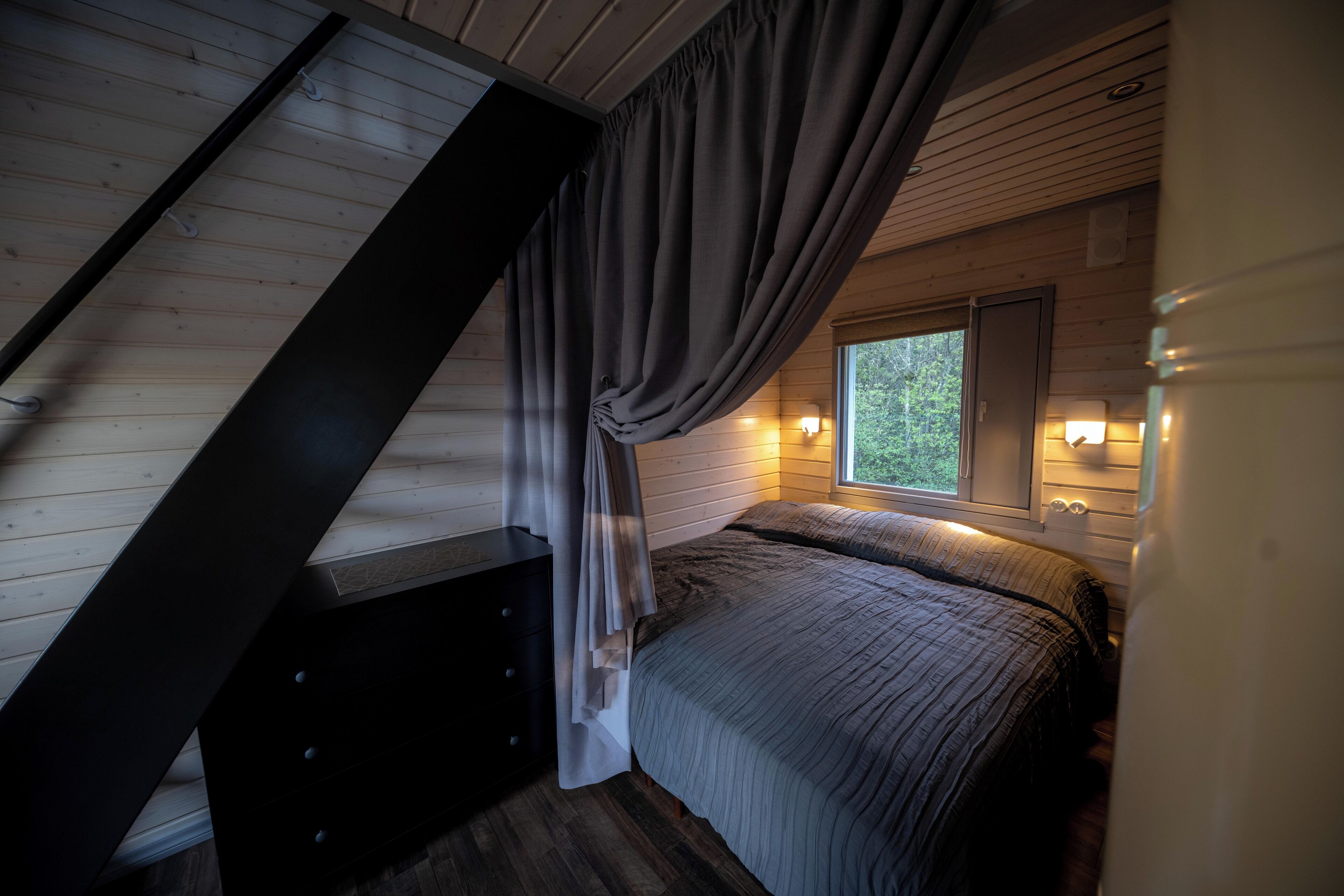A bedroom with a sloped ceiling, a bed with a gray blanket, a nightstand, and a window with a view of greenery.