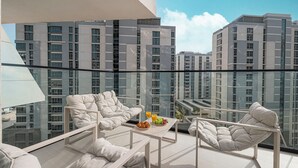 A balcony with two white cushioned chairs, a small table with a fruit plate and a glass of orange juice, and a clear view of surrounding high-rise buildings. - Binghatti Phantom Suites by Hostiva (Dubai)