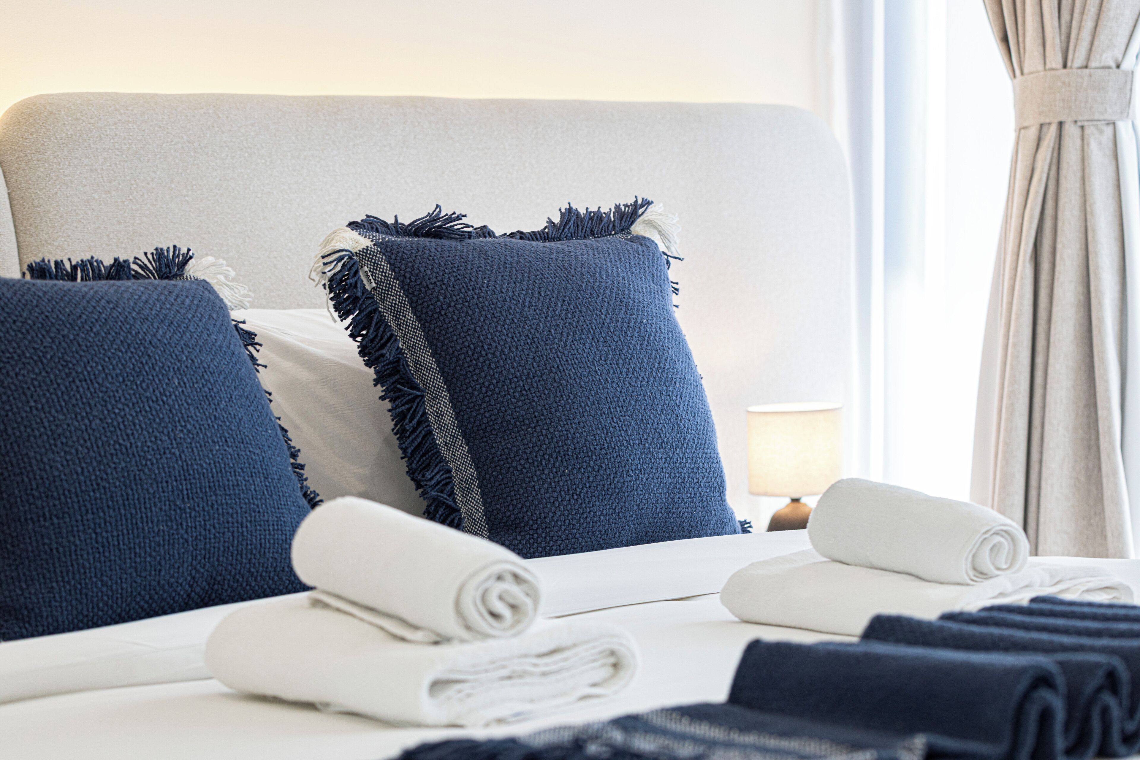 A neatly made bed with rolled white towels and blue pillows with fringes.