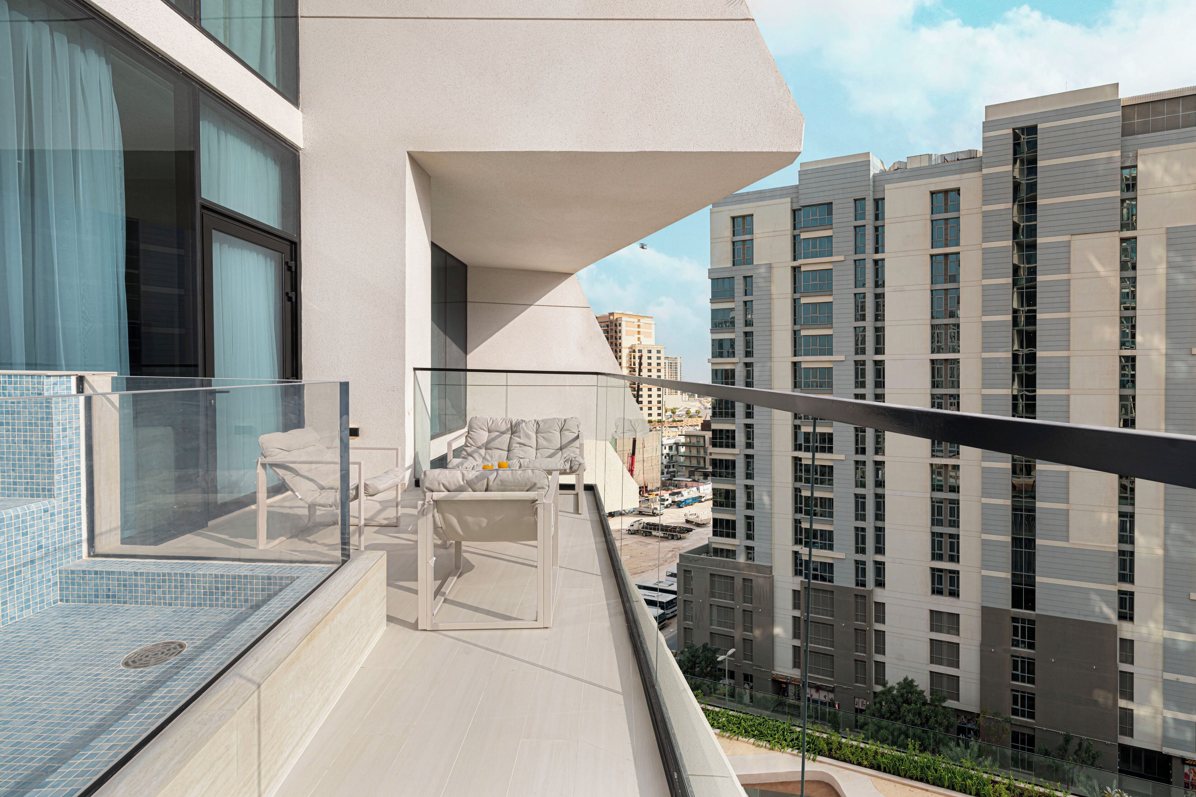 Un balcón moderno con barandilla de vidrio, una zona de estar con cojines y vistas a edificios altos circundantes.