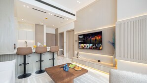 A modern living room with a wooden coffee table, bar stools, a sofa, and a TV mounted on the wall displaying a movie. - Binghatti Phantom Suites by Hostiva (Dubai)