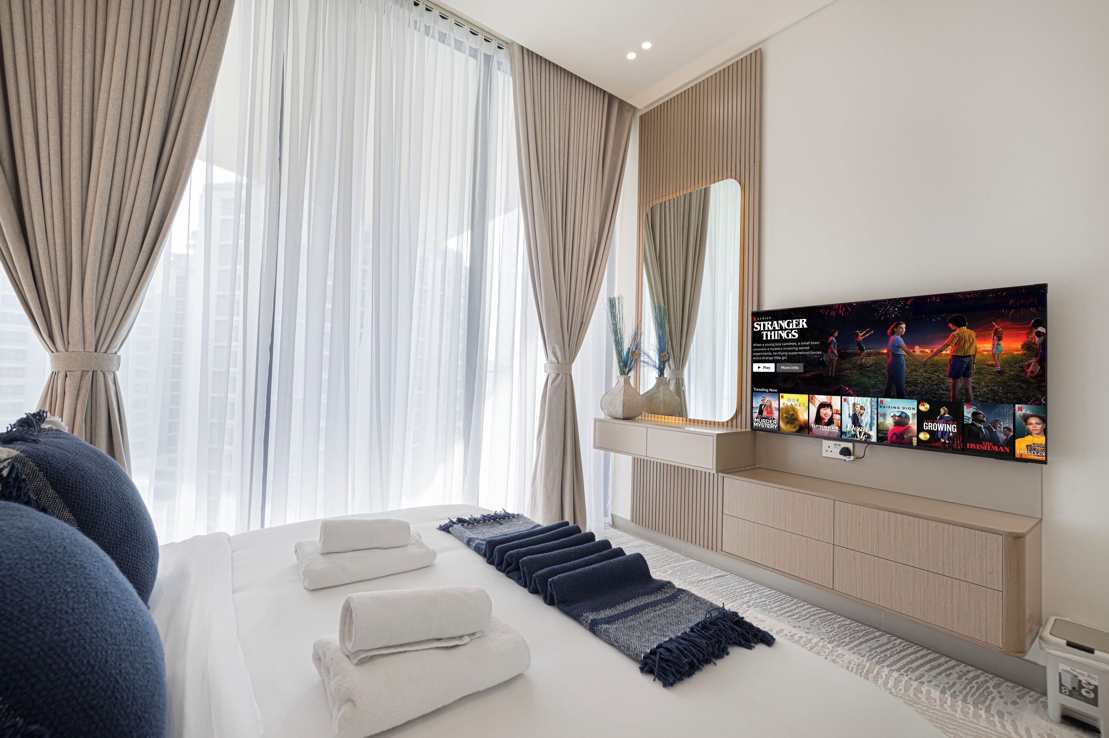 A modern hotel room with a large bed, a flat-screen TV mounted on the wall, and a large window with sheer curtains.