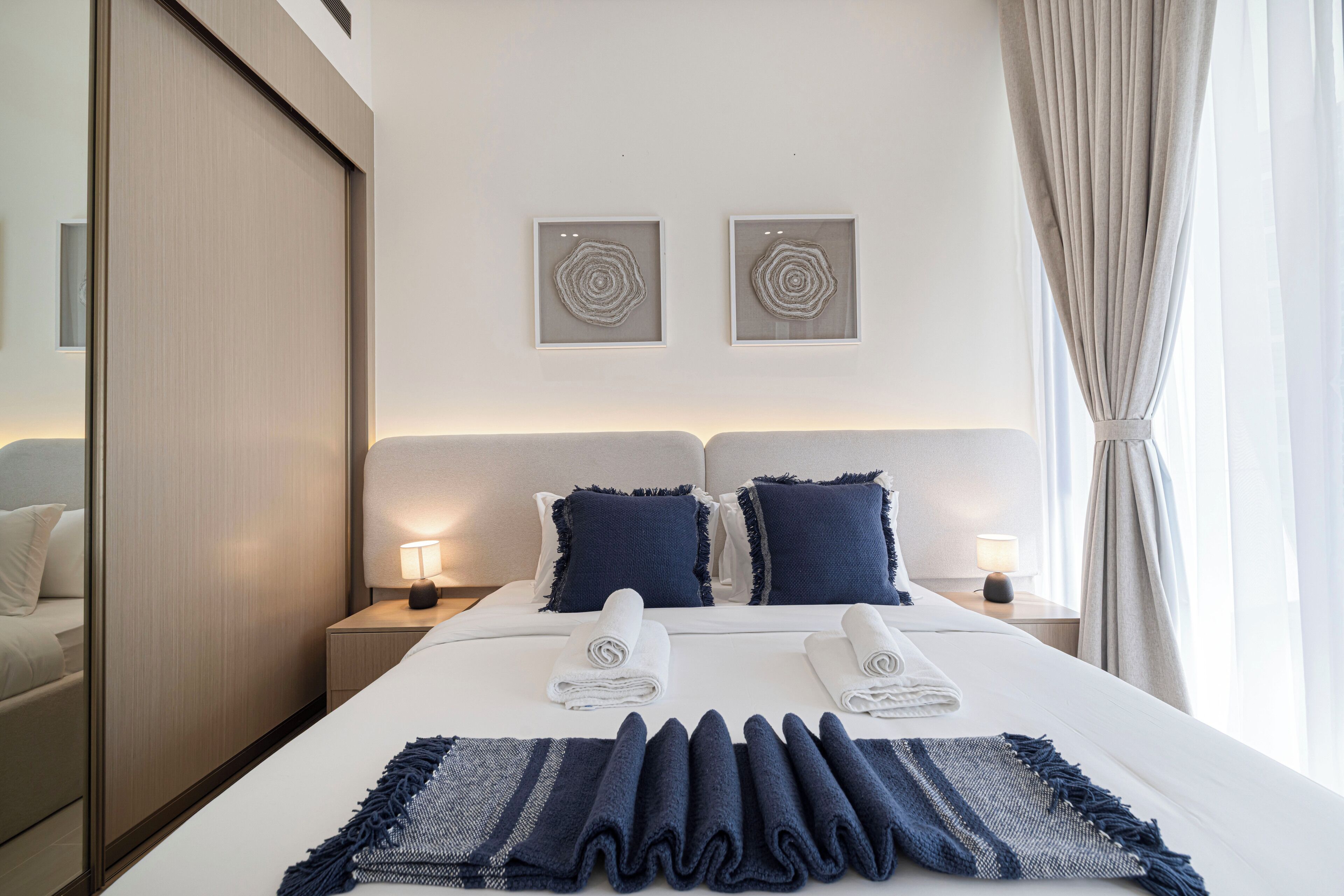 A hotel room with a neatly made bed, two bedside tables with lamps, a large window with curtains, and two wall art pieces.
