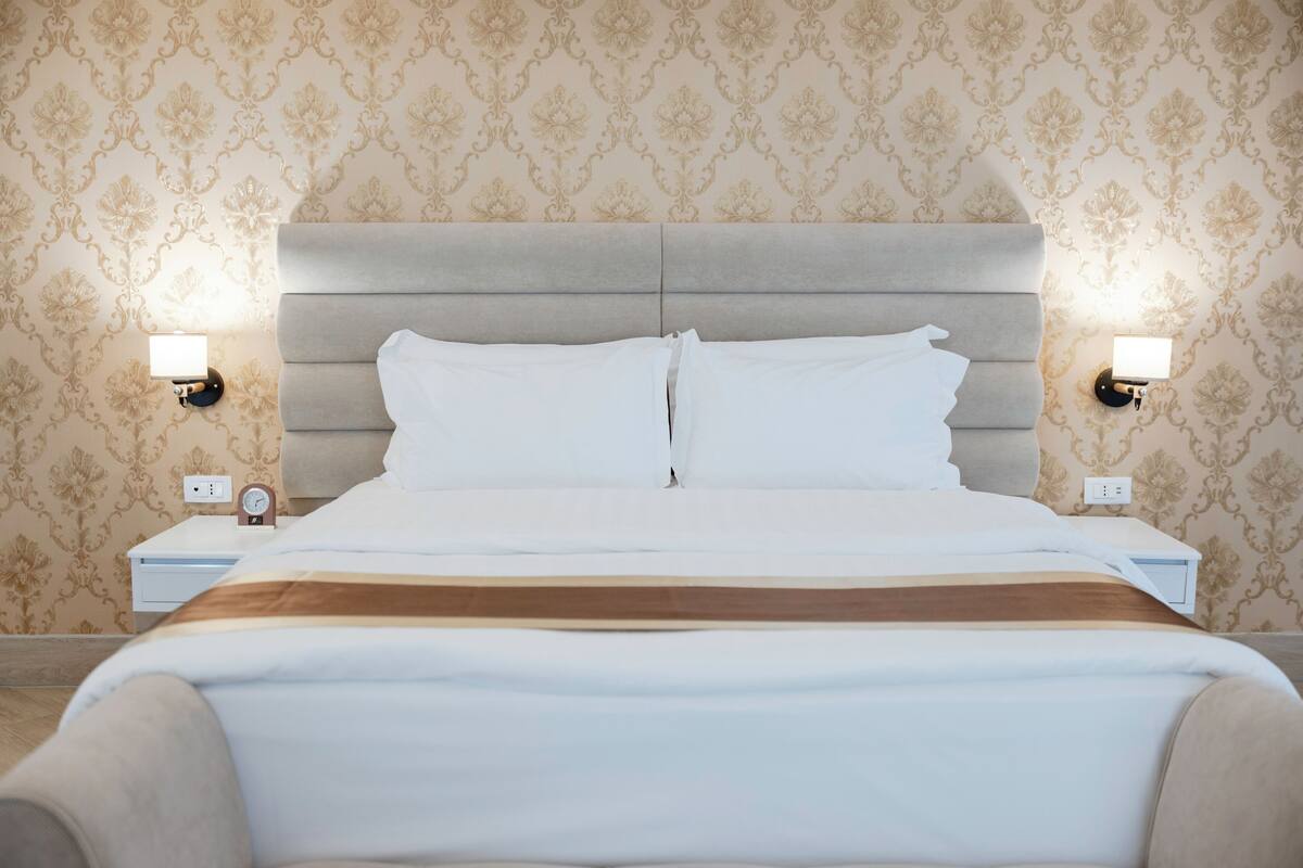 A neatly made bed with white linens and a brown accent, flanked by two bedside tables with lamps, set against a patterned wallpaper.