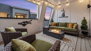 Eine Dachterrasse mit Korbmöbeln, einer Feuerstelle, Lichterketten und Blick auf den Sonnenuntergang.