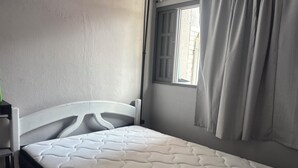 Habitación individual con una cama que tiene cabecera blanca, ventana con cortinas y un colchón con cubierta estampada.