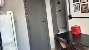 Habitación compacta con puerta gris, un refrigerador blanco, una mesa de comedor con sillas y una mesita con una caja roja.