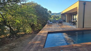 A modern outdoor pool area with a wooden deck, seating, and a building with large windows. - CORSICA: LOUNICELI Villa with air conditioning, a pool, beaches, shops, and mountains (Zonza)