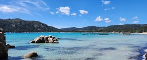 Een kustlandschap met helder turquoise water, rotsformaties en verre bergen onder een blauwe lucht.
