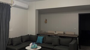 A modern living room with a dark-colored sofa, a small round table with a rug, a ceiling light fixture, and a dining table with chairs. - Peaceful Zayed Rooftop Stay Near Arkan & Park St. (Al Sheikh Zayed City)