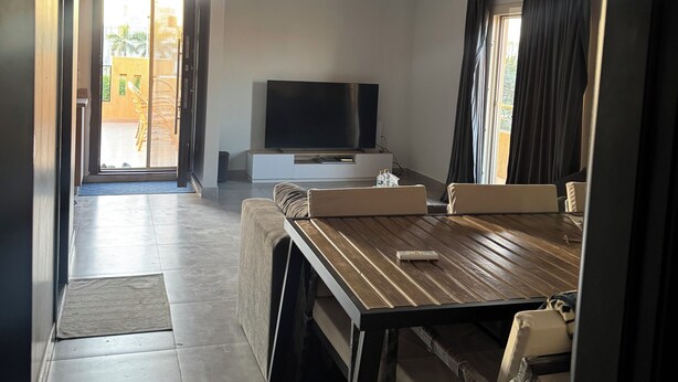 A modern living room with a wooden dining table, a sofa, a flat-screen TV, and a large window with curtains. - Peaceful Zayed Rooftop Stay Near Arkan & Park St. (Al Sheikh Zayed City)