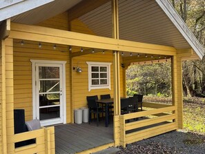 Un bâtiment en bois jaune avec un porche couvert, deux fenêtres, une table et des chaises, et une poubelle.