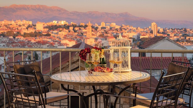 Un balcón con una mesa dispuesta para dos personas, que incluye una botella de vino, copas y un arreglo floral. El balcón tiene vistas a un paisaje urbano con montañas al fondo.
