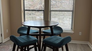 En matplats med ett runt bord och fyra stolar, stora fönster med persienner och en hängande lampa.