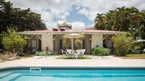 Un área junto a la piscina con piscina, un porche cubierto con mesa y sillas, y un edificio con techo rojo.