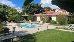 Un área de piscina con sillones de descanso, un pabellón de techo de paja y un edificio con techo rojo.