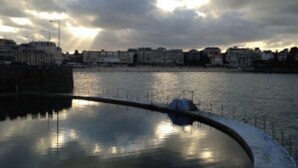 Een uitzicht op het water met een rustig wateroppervlak, een gebogen steiger en gebouwen op de achtergrond onder een bewolkte lucht.