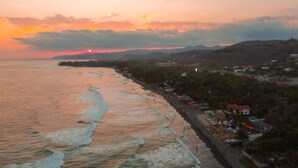A coastal town at sunset with a beach, buildings, and hills in the background.
