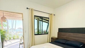 A bedroom with a bed, wooden flooring, a window with curtains, and a view of the outdoors.
