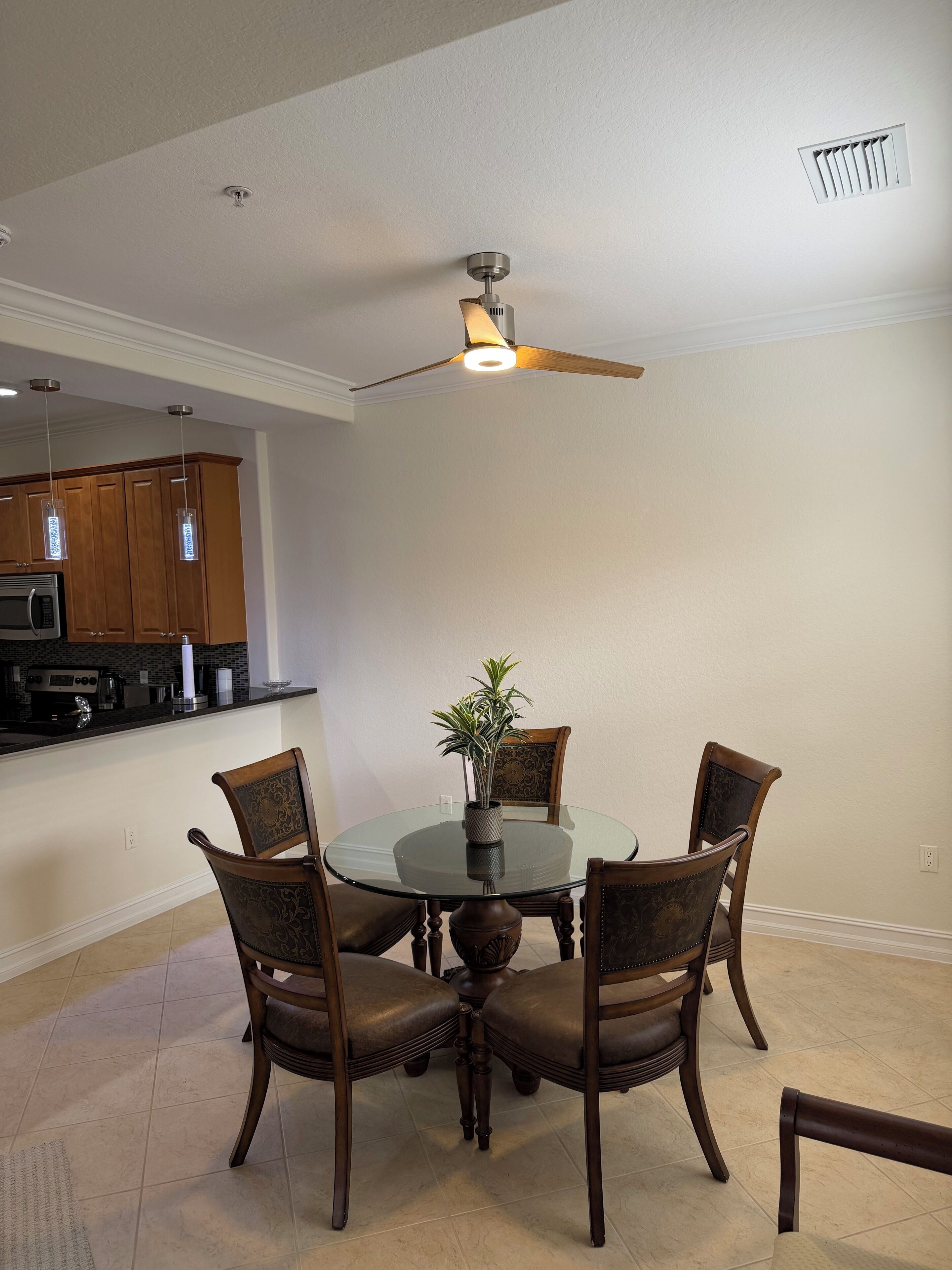 A dining area with a glass table surrounded by wooden chairs, a ceiling fan, and a kitchen in the background.