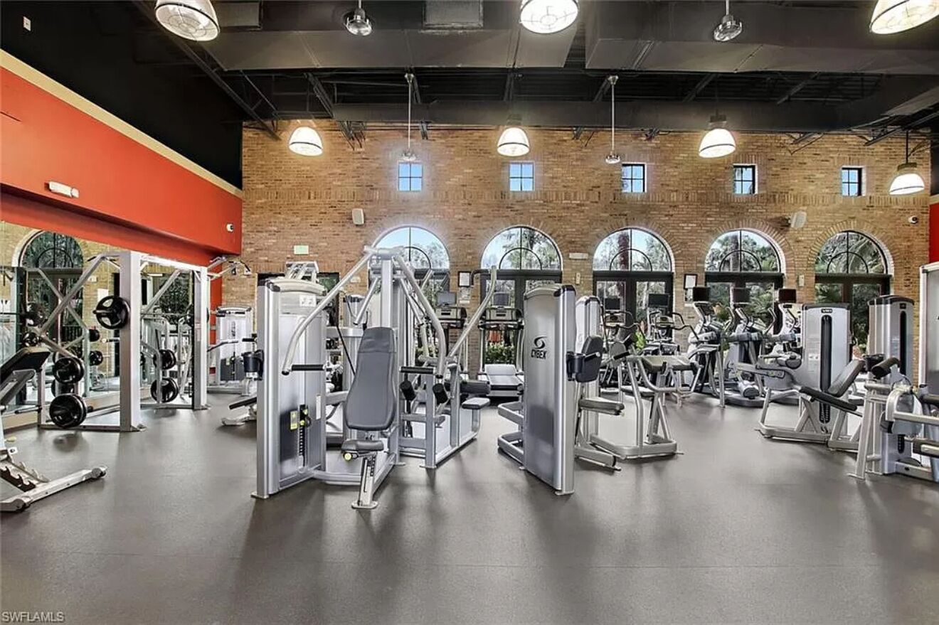 Une salle de sport équipée de divers appareils d’exercice, dotée de grandes fenêtres et d’un mur en briques.