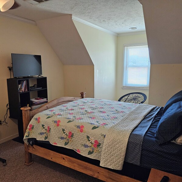 A bedroom with a sloped ceiling, a bed with a floral quilt, a television on a stand, and a window with blinds.