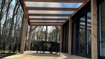 A wooden deck with a glass ceiling, a table with chairs, and a view of trees.