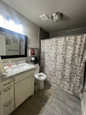 Un baño con lavamanos blanco, espejo y váter. Hay una cortina de ducha con estampado de hojas.