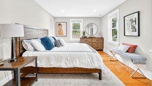 A modern bedroom with a large bed, bedside table, lamp, dresser, bench, and artwork. - Charming Renovated Home, Cozy for All! Pet friendly! (New Windsor)
