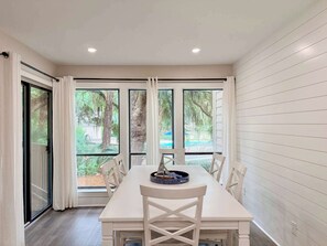 A dining area with a long table, white chairs, and a view of trees and a pool through large windows. - Coastal Retreat with Private Balcony & Pool Access (Hilton Head Island)