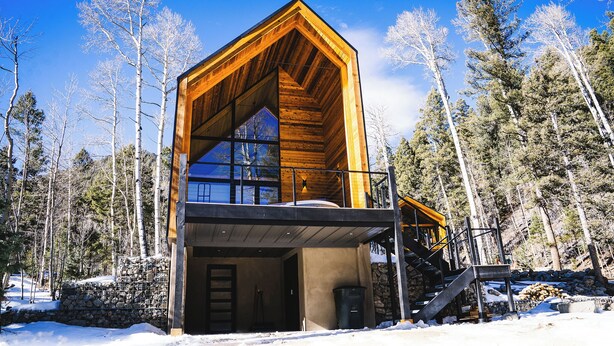 A modern log cabin with a large balcony and a stone foundation, surrounded by a snowy forest.