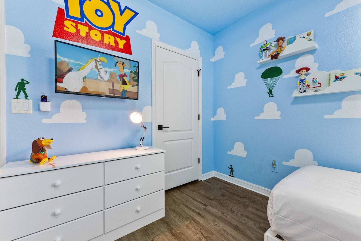 A room with a blue wall, a white door, a white dresser, and a bed. There are toys and a TV on the wall.
