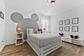Un dormitorio con cama, mesita de noche, lámpara, decoración de Mickey Mouse en la pared y cuadros enmarcados.