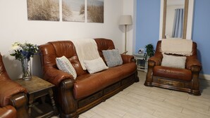 Ein Wohnzimmer mit braunem Ledersofa, einem passenden Sessel, einem kleinen Holztisch und einer Vase mit Blumen.
