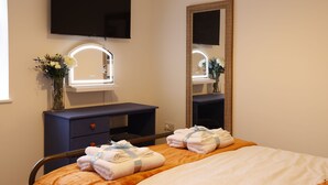 Ein Hotelzimmer mit einem Bett, einem Schminktisch mit Spiegel, einem an der Wand montierten Fernseher und einem Fenster mit Blumen.