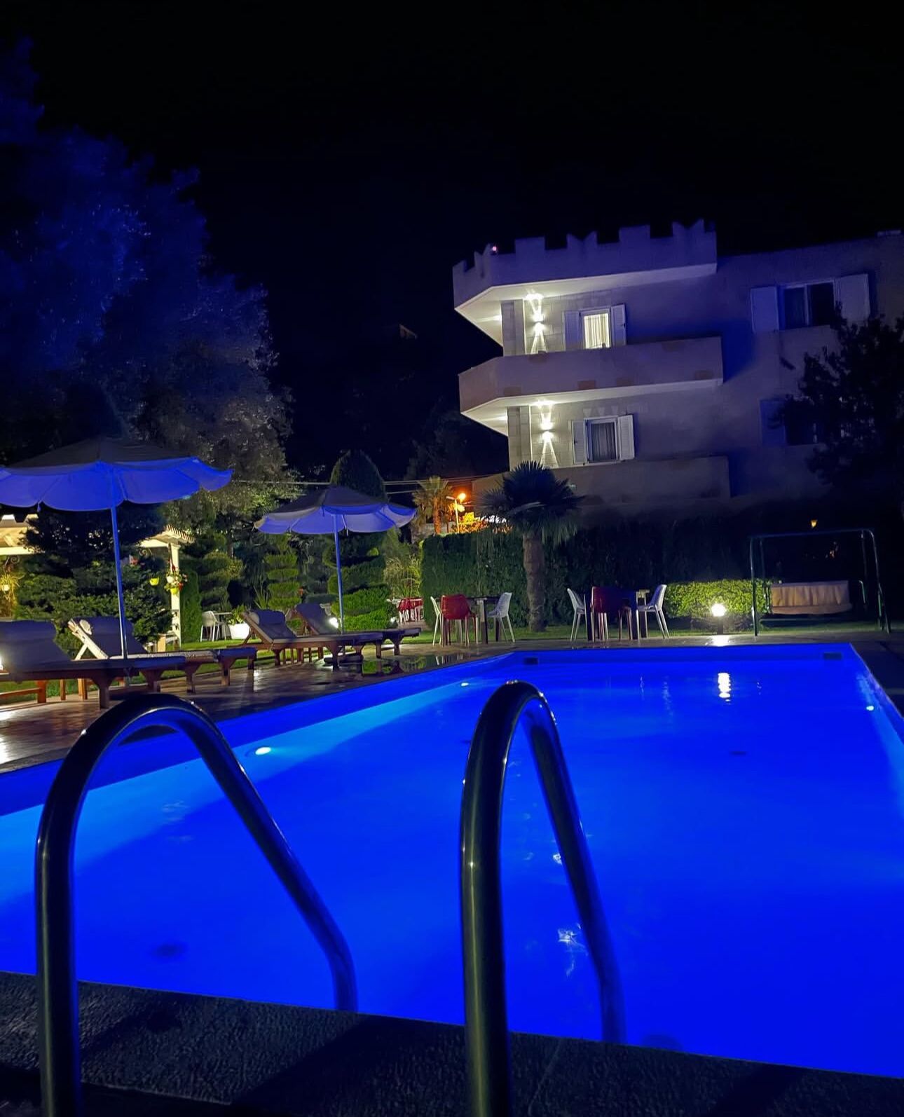 A swimming pool with a blue tint, surrounded by lounge chairs and umbrellas, with a multi-story building in the background.