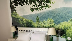 Een slaapkamer met een bed, twee nachtkastjes, twee lampen, een witte spreidek en een muurschildering van een bos en bergen.