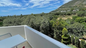 Een balkon met een witte tafel en rode stoel, uitkijkend op een bergachtig landschap met weelderige begroeiing en een helderblauwe lucht.