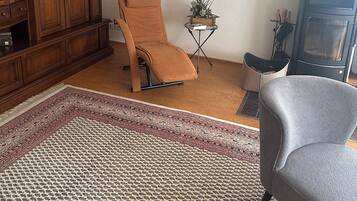 Ein Wohnzimmer mit einem Holzkasten, einem braunen Sessel, einem grauen Sessel, einem gemusterten Teppich und einem kleinen Tisch mit einer Pflanze.