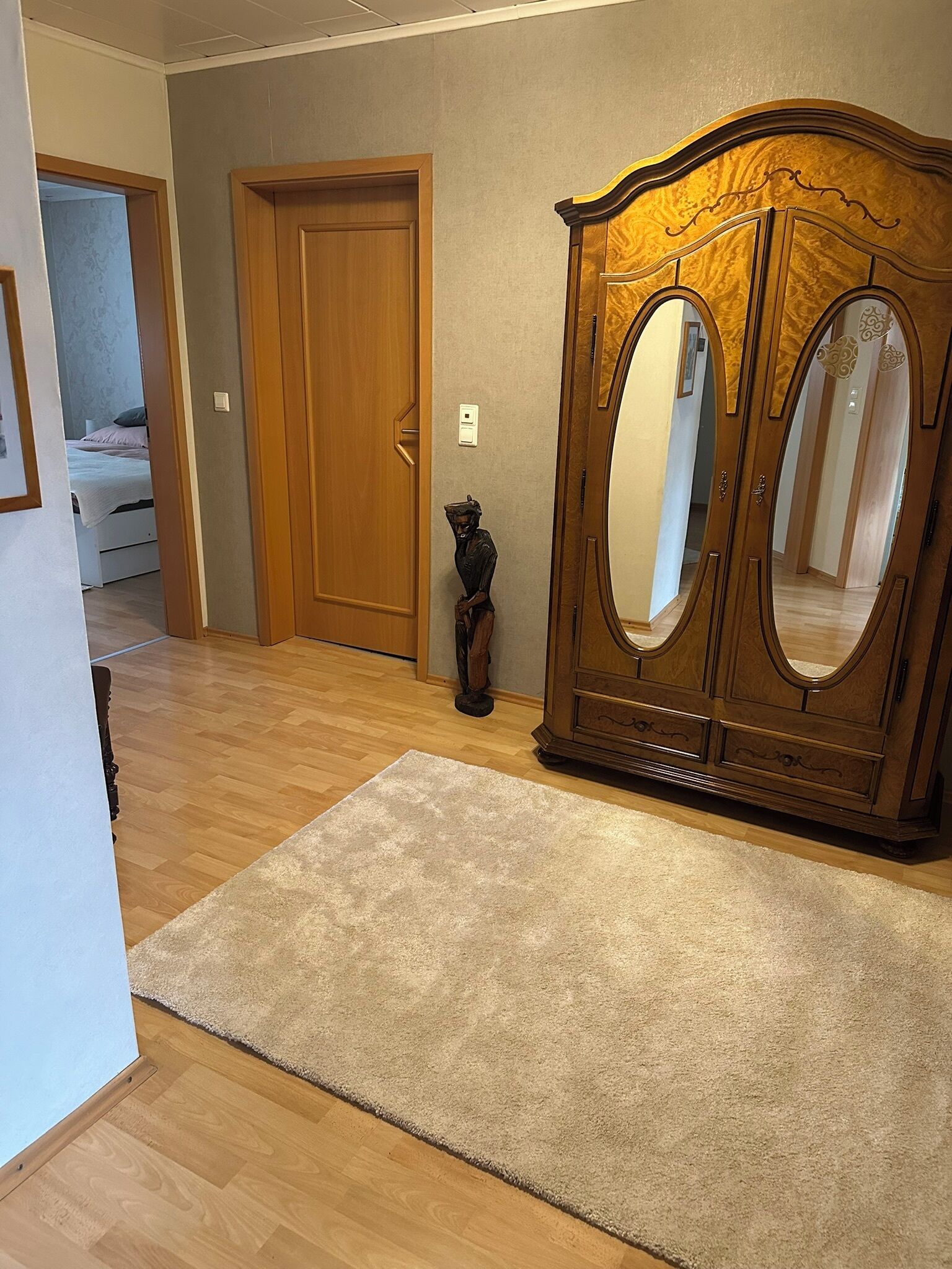 A hallway with a wooden floor, a beige rug, a wooden wardrobe with two doors, and a small statue.