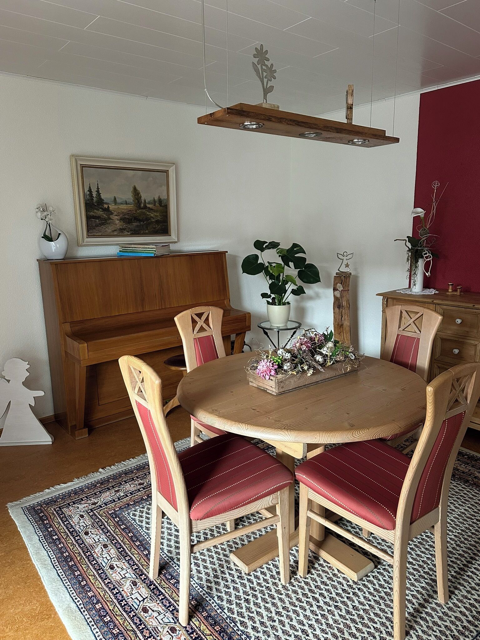 A dining area with a round wooden table, red upholstered chairs, a wooden piano, and a painting on the wall.