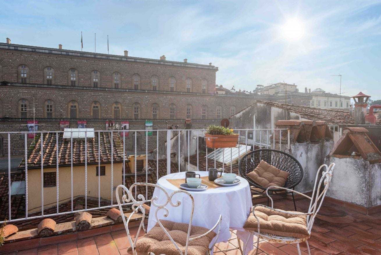 A rooftop terrace with a round table, white metal chairs, and a view of a historic building.