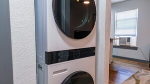 A compact, modern laundry room with a front-loading washer and dryer, wooden flooring, and a window with blinds. - Cozy retreat minutes from AT&T Stadium (Arlington)