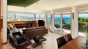 Ein geräumiger Wohnbereich mit großem Fenster und Blick auf die Stadt, ein Balkon mit Meerblick und eine Essecke mit Tisch und Stühlen.