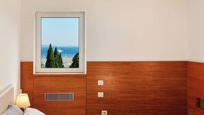 Ein Schlafzimmer mit Holz-Kopfteil, einem Fenster mit Meerblick, einem Bett mit grau-weißer Bettwäsche und einem Nachttisch mit Lampe.