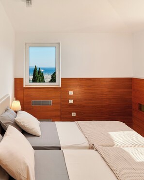 Ein Schlafzimmer mit Holz-Kopfteil, einem Fenster mit Meerblick, einem Bett mit grau-weißer Bettwäsche und einem Nachttisch mit Lampe.