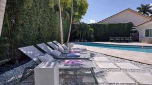 Un espace aménagé au bord de la piscine, avec des chaises longues et une piscine, le tout entouré de verdure et d’un bâtiment.