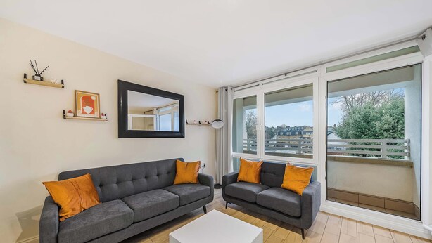 Un salon moderne avec deux canapés gris, une table basse blanche et une grande fenêtre donnant sur des immeubles et des arbres.