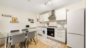Une cuisine moderne avec des armoires blanches, une hotte de cuisinière en acier inoxydable, un four encastré et un réfrigérateur. On y trouve une table à manger avec des chaises et un tapis sur le sol.